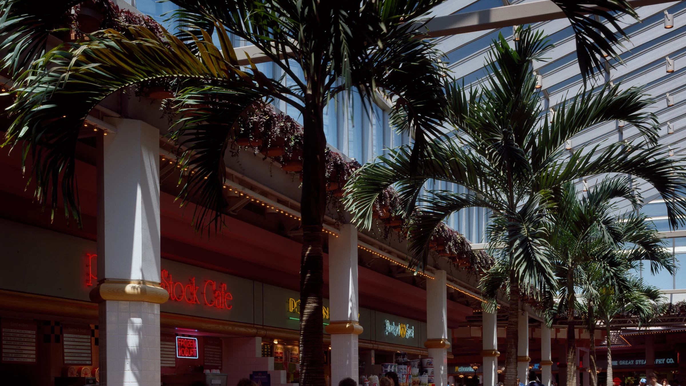 Shoppers walking through a bright 1980s indoor mall beneath a glass roof, with palm trees and food counters lining the tiled concourse.